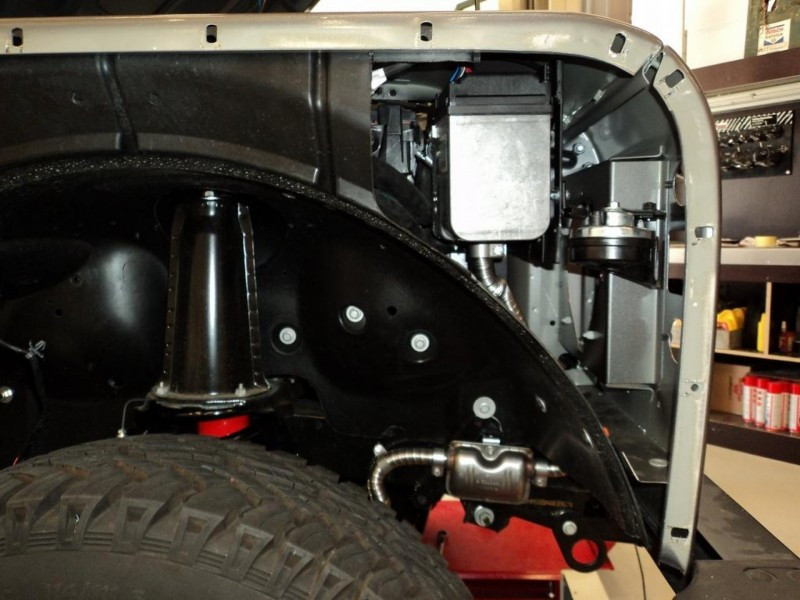 Vehicle wheel well showing suspension components and exposed inner frame with wiring and hoses.