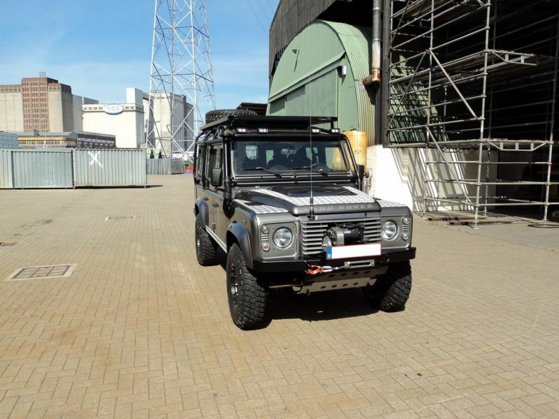 Front view of a silver/gray Land Rover Defender parked on a paved industrial yard with scaffolding and large green hangar in the background.