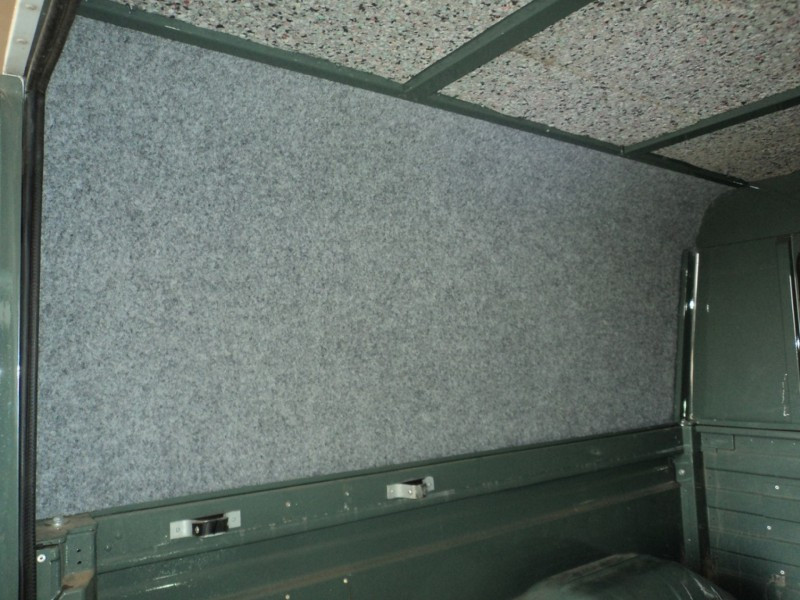 Interior of a cargo van with gray speckled carpeting on the walls, green metal framing, and a partially visible spare tire at the bottom right.