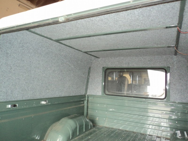 Interior of a pickup truck bed lined with grey carpeting, small side window visible on the right side, green metal panels.