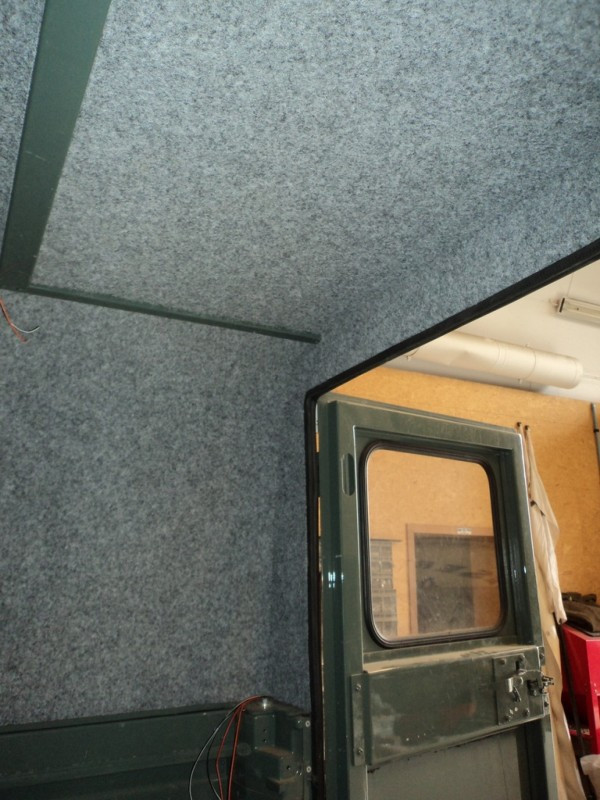 Interior of a blue-carpeted room with a green metal door ajar, revealing a window and a workshop area behind it