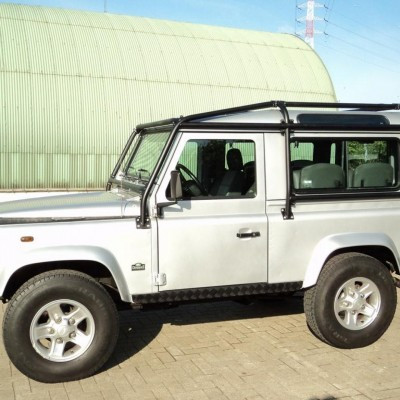 Silver off-road SUV with a roof rack parked on a brick lot, a large green curved building in the background.