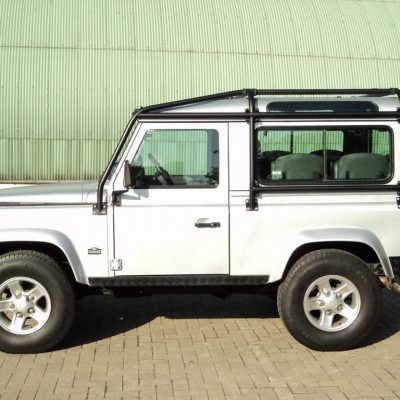 Side view of a white 4x4 SUV with a roof rack parked on a brick surface, green corrugated metal wall in the background.