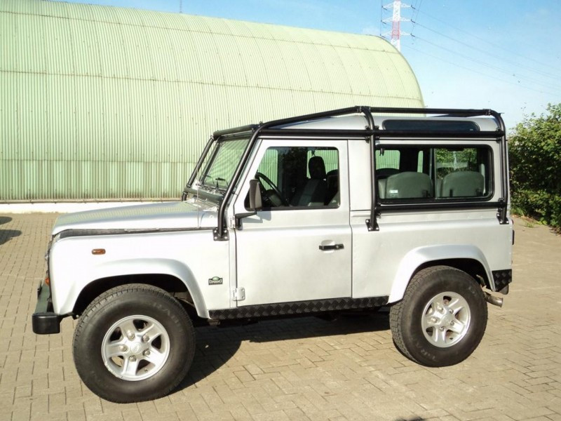Silver Land Rover Defender SUV with a roof rack parked on a brick lot, green curved industrial building in the background