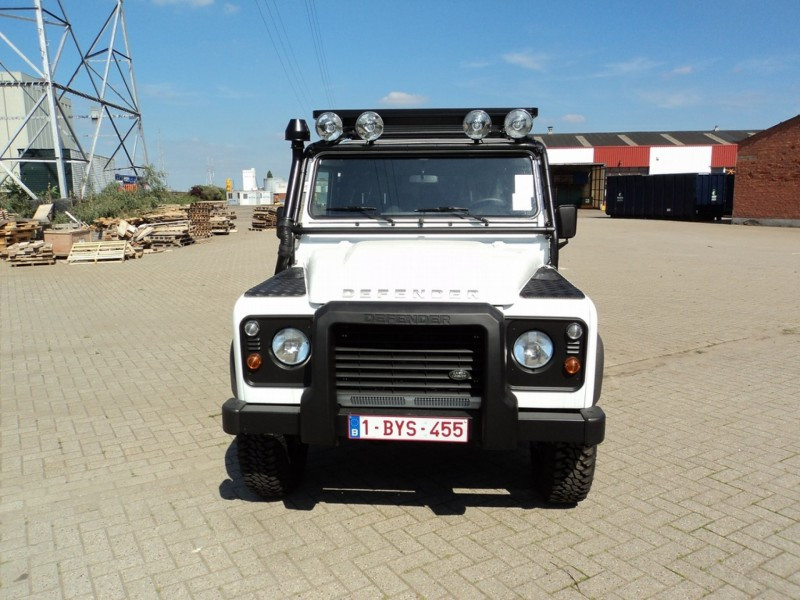 Front view of a white Land Rover Defender with a roof light bar, parked on a brick yard with pallets and industrial buildings in the background.