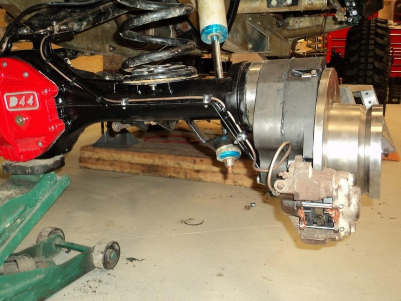 Front suspension and braking assembly of a lifted vehicle: D44 axle, coil spring, blue shocks, large rotor and rusty caliper visible in a workshop.