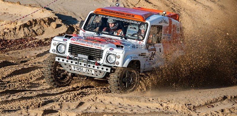 White off-road rally truck speeding through a sandy track, splashing mud as it kicks up dust behind it.