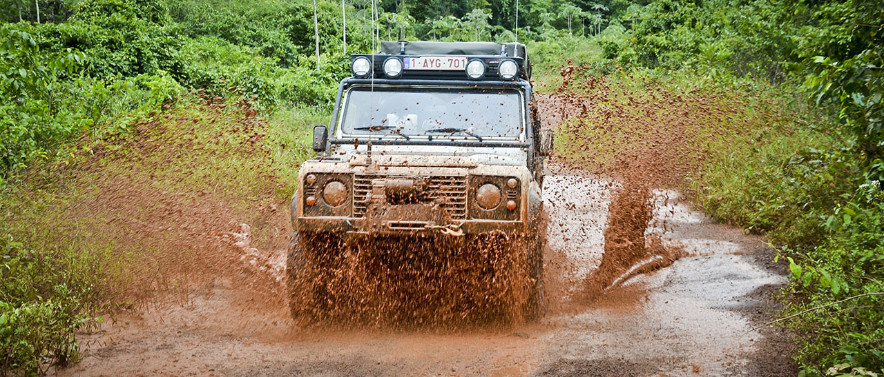 White off-road SUV driving through a muddy red puddle on a forest trail, mud spraying up around the vehicle's sides.