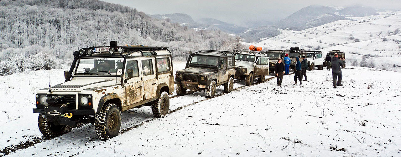 Row of muddy 4x4 off-road vehicles lined up on a snowy hillside with people nearby.